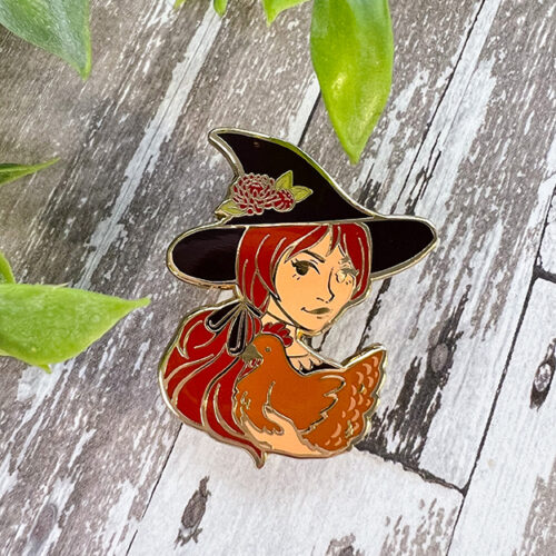The Haunted Cottagecore ~ Enamel Pin features a red-haired witch in a black hat with a flower, holding a brown chicken, displayed on weathered wood with green leaves in the corners.