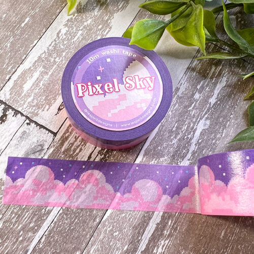 A roll of Pixel Sky Washi Tape rests on a wooden surface with nearby plants. The tape displays a pixel art design featuring pink clouds and stars set against a purple sky.