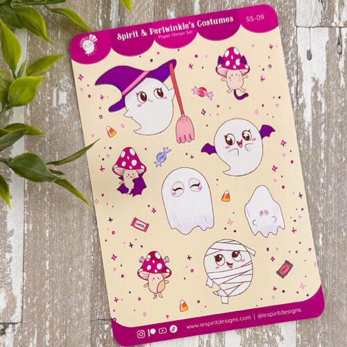 The "Spirit & Periwinkle's Costumes ~ Paper Sticker Sheet" features cute cartoon ghosts, a cat, mushrooms, and candies in Halloween costumes on a pastel sheet with a pink border, displayed on wood beside green leaves.