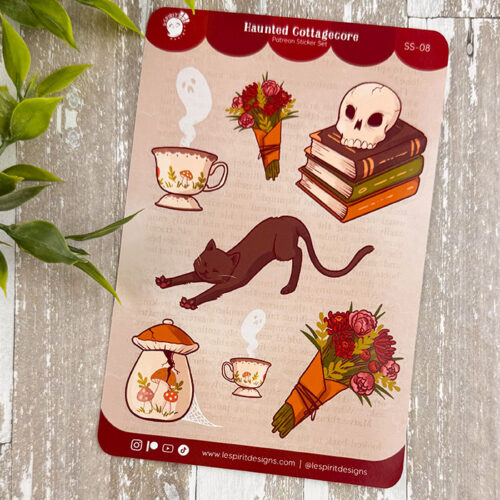 The Haunted Cottagecore ~ Paper Sticker Sheet showcases whimsical designs—black cat stretching, skull on books, flowers, ghostly teacups, and potion bottles—on a wooden background with green leaves nearby.