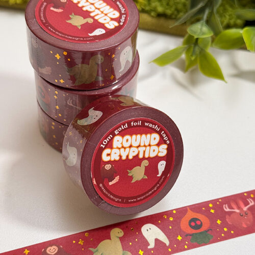 Four rolls of Round Cryptids Foil Washi Tape are stacked next to a plant. The tape shows cute, round cryptid designs—ghosts, Bigfoot, and stars—on a red background. A single strip is unrolled on the table.