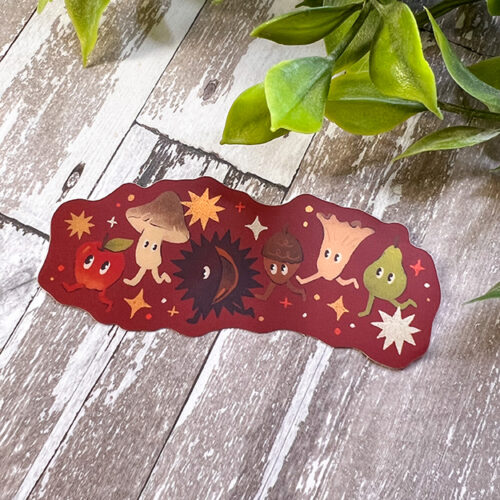 A colorful sticker with cute cartoon characters, including an apple, mushroom, sunflower, acorn, tree stump, and leaf, surrounded by stars, placed on a wooden surface near green leaves.