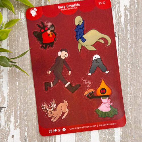 The Cozy Cryptids ~ Paper Sticker Sheet features six vibrant cartoon cryptids in cozy outfits on a red background, with a leafy plant in the top left and a rustic wooden surface underneath.