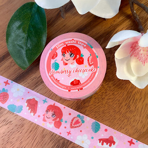 A roll of pink washi tape labeled strawberry cheesecake with a cartoon girl, strawberries, and cheesecake illustrations, shown on a wooden surface next to white flowers.
