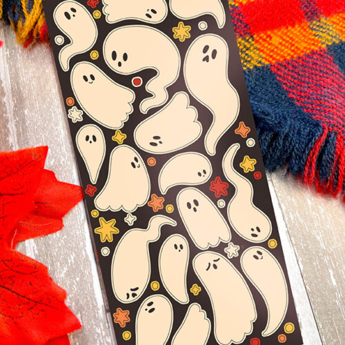 The Silly Spooky Ghosts ~ Deco Sticker Sheet showcases cute cartoon ghosts with fun expressions, accented by stars and dots. Displayed on a wooden surface, it's placed beside a red leaf and colorful plaid fabric.