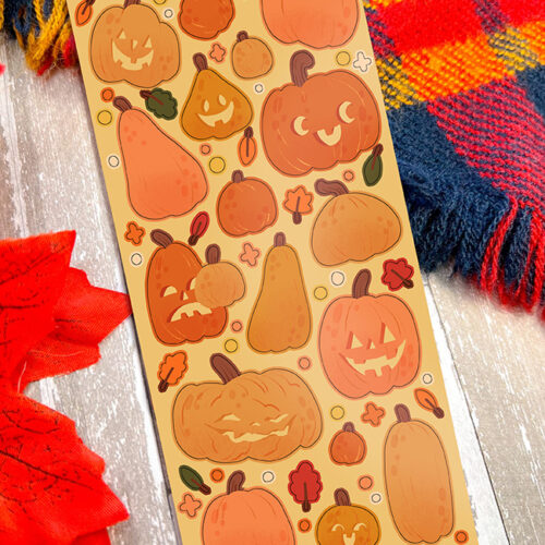 A rectangular Silly Spooky Ghosts ~ Deco Sticker Sheet featuring playful ghost designs, displayed on a rustic wooden surface next to a plaid blanket and an orange decorative leaf.
