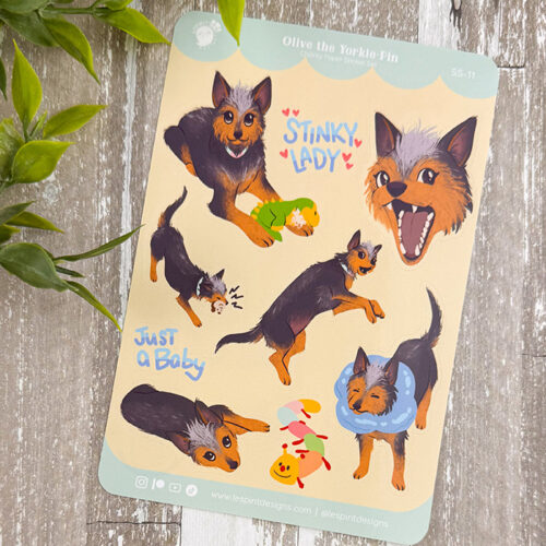 The Olive the Yorkie-Pin ~ Vet Charity Sticker Sheet features playful Yorkshire Terrier stickers with phrases like STINKY LADY and JUST A BABY, displayed on a rustic wooden background with green leaves.