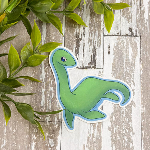 The Nessie ~ 3" Matte Vinyl Sticker, featuring a cute green cartoon dinosaur with a long neck and tail, is displayed on a rustic white wooden surface with green leafy branches in the top left corner.