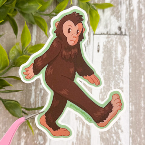 The Bigfoot ~ 3" Matte Vinyl Sticker shows a cartoon Bigfoot with brown fur walking on two legs, one arm forward and one back. It's held by tweezers over green leaves on a light wooden surface.