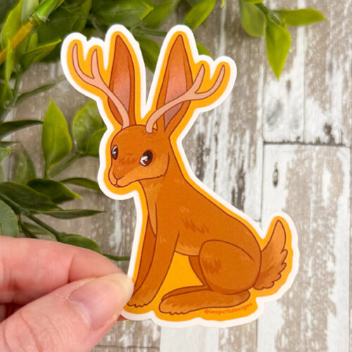 A hand holds the Jackalope ~ 3" Matte Vinyl Sticker, featuring a brown mythical rabbit with antlers, set against green leaves and wooden planks.