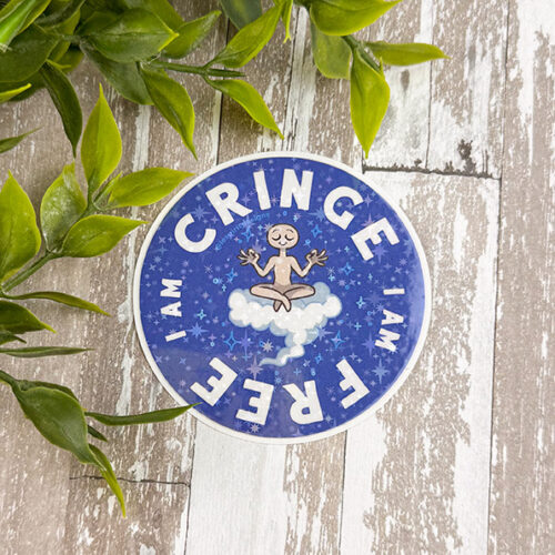 The 3" Sparkle Holo Vinyl Sticker, "I Am Cringe," features a meditating cartoon figure on a cloud with stars, surrounded by the text "I AM CRINGE I AM FREE." Green leaves appear in the corner against the wooden background.