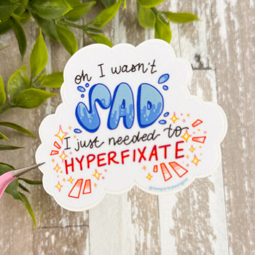 The Hyperfixate ~ 3" Matte Vinyl Sticker features a white cloud with "oh I wasn't SAD, I just needed to HYPERFIXATE"—SAD in blue bubble letters and HYPERFIXATE in bold red—plus colorful doodles and green leaves around the design.