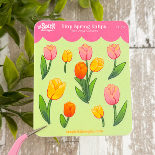 The Tiny Tulips ~ Tiny Filler Vinyl Sticker Sheet displays colorful vinyl stickers of small pink and orange tulips with green leaves. Green leaves and wooden boards are partially visible in the background.