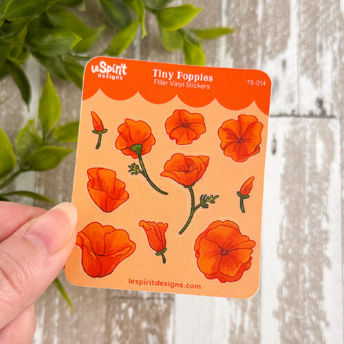 A hand holds the Tiny Poppies ~ Tiny Filler Vinyl Sticker Sheet, featuring small orange poppy illustrations on an orange background, with green leaves and a rustic wooden surface behind.