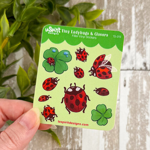 A hand holds the Tiny Ladybugs & Clovers ~ Tiny Filler Vinyl Sticker Sheet, featuring red ladybugs—some flying, some crawling—and green clovers on a light green background.