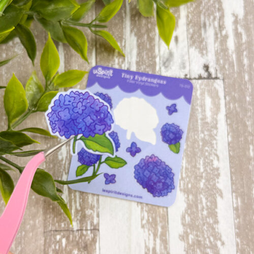 A hand uses pink tweezers to place a sticker from the Tiny Hydrangeas ~ Tiny Filler Vinyl Sticker Sheet onto a sheet with blue and purple hydrangea designs, near green artificial leaves on a wooden surface.