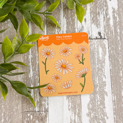 The Tiny Daisies ~ Tiny Filler Vinyl Sticker Sheet features small white daisies with yellow centers on an orange background, plus green leaves on the left, displayed atop a wood-patterned surface.