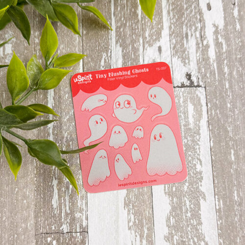 The “Tiny Blushing Ghosts ~ Tiny Filler Vinyl Sticker Sheet” features nine cute white cartoon ghosts on a pink background with green leaves, displayed on a weathered wooden surface.