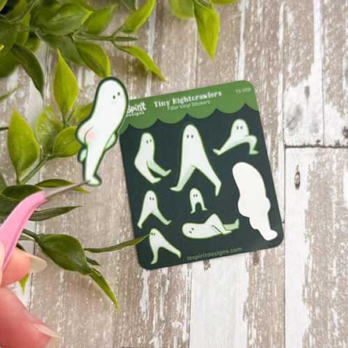 A hand holds a small sticker from the Tiny Nightcrawlers ~ Tiny Filler Vinyl Sticker Sheet, featuring a cute white ghost with green outlines near matching stickers on rustic wood, with green leaves in the corner.