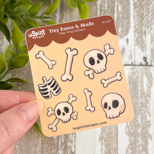 A hand holds the Tiny Bones & Skulls ~ Tiny Filler Vinyl Sticker Sheet, featuring cartoon skulls, bones, and a ribcage. A blurred green plant appears in the background.