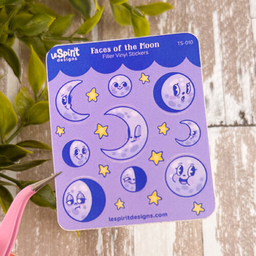 The Tiny Faces of the Moon ~ Tiny Filler Vinyl Sticker Sheet features cartoon moons and stars with expressive faces on a purple background, surrounded by green leaves and displayed on a wooden surface.