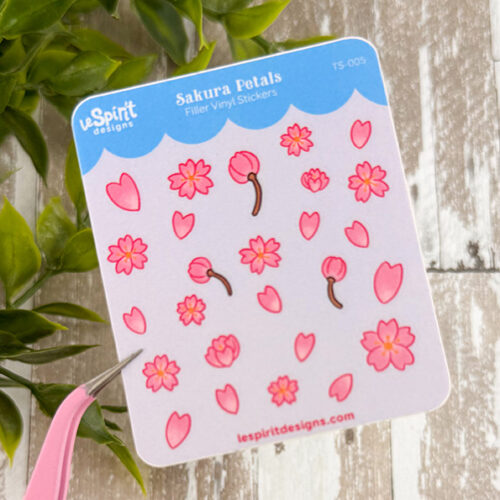 A Tiny Sakura Petals ~ Tiny Filler Vinyl Sticker Sheet with pink cherry blossom flowers and petals is held by tweezers against a leafy background on a wooden surface.