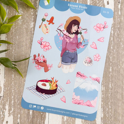 The Hanami Picnic ~ Paper Sticker Sheet is displayed on a wooden surface, showcasing Japanese designs with a girl, cherry blossoms, food, Mt. Fuji, picnic items, and a green plant partly visible in the upper left corner.