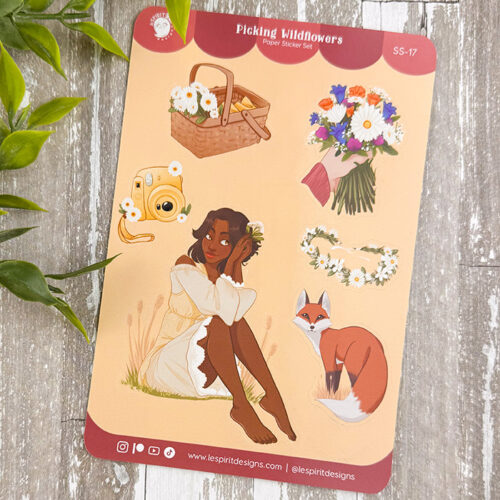 The Picking Wildflowers ~ Paper Sticker Sheet features a girl in a white dress and flower crown, a fox, bouquet, basket, camera, and flower crowns on a wooden surface with green leaves in the corner.