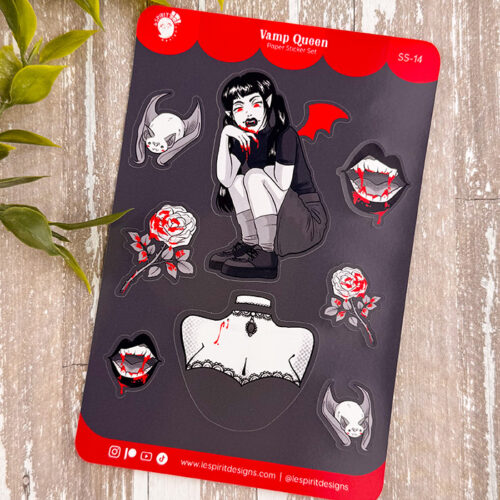 The Vamp Queen ~ Paper Sticker Sheet showcases a gothic girl with bat wings, vampire bats, fanged mouths, roses, and a laced corset on a dark background, displayed on wood with green leaves nearby.