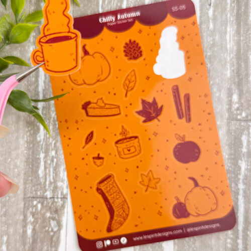 A hand holds a sticker of a steaming mug above an orange autumn-themed sticker sheet featuring pumpkins, leaves, a candle, a sock, a pinecone, and other fall items on a wooden surface with green leaves nearby.