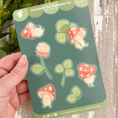 A hand holds a sheet of stickers featuring cute mushrooms with faces, a pink clover flower, and clover leaves. The sheet is labeled Periwinkle the Mushroom Boi on a green background.