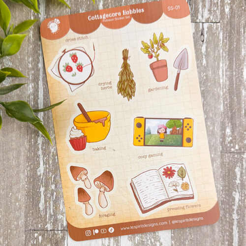 A sticker sheet titled Cottagecore Hobbies featuring illustrations of cross-stitch, drying herbs, gardening, baking, cozy gaming, foraging mushrooms, and pressing flowers, on a wood surface with green leaves.