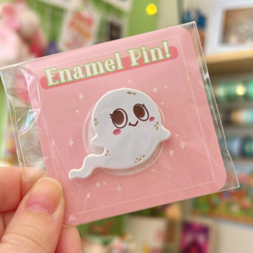 A hand holds the Spirit ~ Dyed Enamel Pin, shaped like a smiling white ghost with big eyes and blushing cheeks, shown on pink backing labeled "Enamel Pin!" in green letters. The background features a colorful shop display.