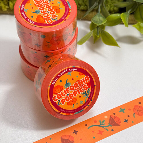 A stack of California Poppies Washi Tape rolls sits on a white surface with greenery behind. A strip featuring poppy flowers and blue dots is displayed in front.