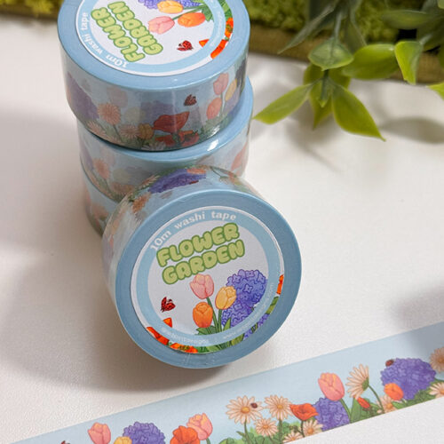 Three rolls of Flower Garden Washi Tape are stacked beside a strip showing colorful flowers and butterflies. Green leaves and moss are in the background.