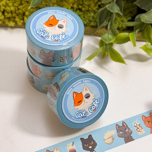 Cat Cafe Washi Tape: Set of three blue rolls featuring playful cartoon cats, coffee cups, pastries, and café-themed designs. One roll is unrolled to display the pattern. Green plants appear in the background.