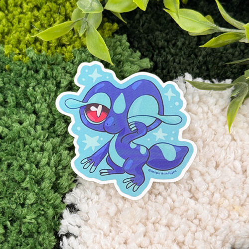 The Uppies Hamster ~ 3" Matte Vinyl Sticker features a cute blue cartoon lizard with big red eyes and a heart-shaped pupil, standing on a green and white textured rug with leafy plants above.