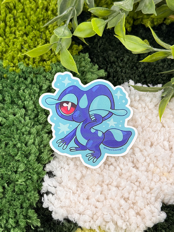 The Uppies Hamster ~ 3" Matte Vinyl Sticker features a cute blue cartoon lizard with big red eyes and a heart-shaped pupil, standing on a green and white textured rug with leafy plants above.