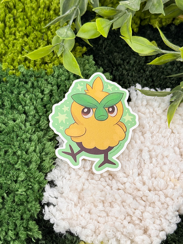 An Uppies Kitty ~ 3" Matte Vinyl Sticker featuring a cute, cartoon cat is displayed on a textured, multicolored rug with green leaves nearby.