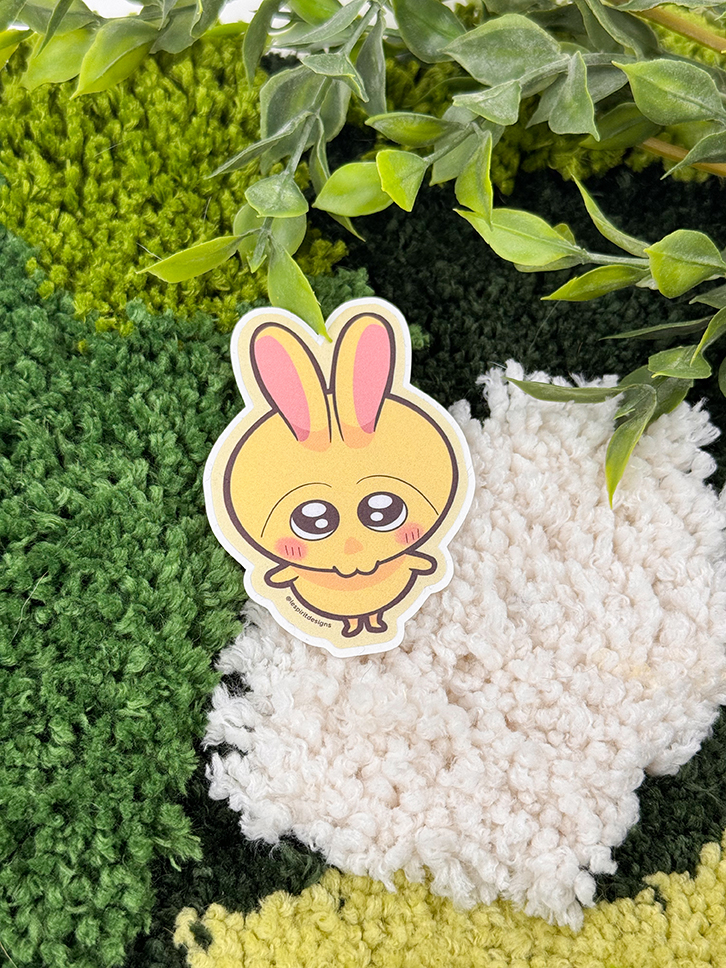 The Uppies Kitty ~ 3" Matte Vinyl Sticker, featuring a cute cartoon cat with big, shiny eyes, is placed on a textured green and white rug with leafy plant stems above it.