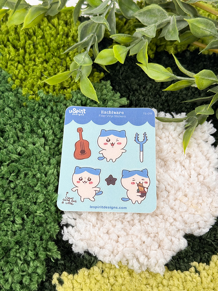 The Tiny Blue Kitty ~ Tiny Filler Vinyl Sticker Sheet features three cute blue and white cat characters, a guitar, and a trident, shown on green and white textured fabric with leafy branches overhead.