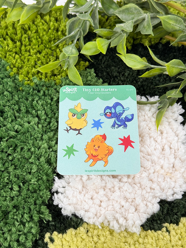 The Tiny Gen10 Starters ~ Tiny Filler Vinyl Sticker Sheet features three colorful cartoon creatures and three stars on a green and white textured fabric, with leafy plants partially covering the top.