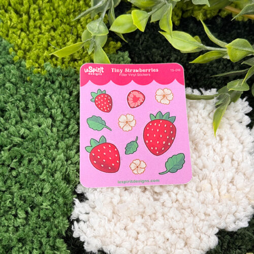 The Tiny Strawberries ~ Tiny Filler Vinyl Sticker Sheet, featuring pink card packaging and strawberry-themed stickers, is displayed on a green and white textured surface with leafy plant stems above.