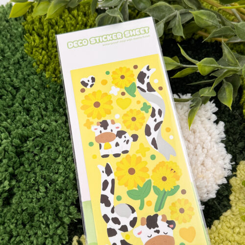 The Sunflower Cows ~ Deco Sticker Sheet features cute cartoon cows, yellow sunflowers, hearts, and green leaves, perfect for decorating journals and crafts. Product is pictured on a green rug with leafy plants in the background.