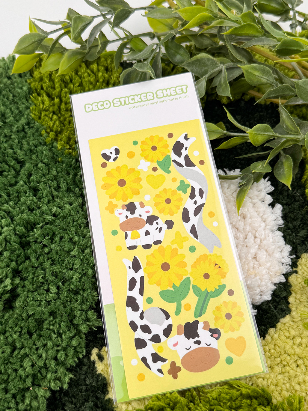 The Sunflower Cows ~ Deco Sticker Sheet features cute cartoon cows, yellow sunflowers, hearts, and green leaves, perfect for decorating journals and crafts. Product is pictured on a green rug with leafy plants in the background.