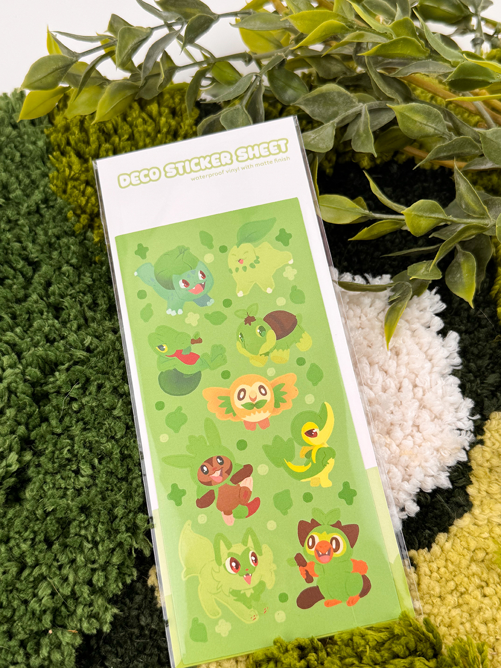 The Grass Starters ~ Deco Sticker Sheet features cute, cartoon-inspired green Pokémon on a leafy background, shown on a textured green and cream rug with artificial plant leaves nearby.