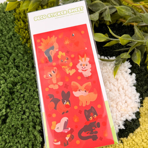 The "Fire Starters ~ Deco Sticker Sheet" features cute, cartoon fire-themed animals on a red background, displayed on a green and cream textured surface with leafy plants nearby.