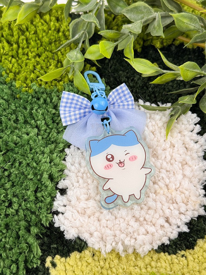 The Blue Kitty ~ Acrylic Keychain features a cute cat-shaped design in blue and white, accented with a small blue bell and gingham bow, displayed on a textured green-and-white rug beside leafy plants.