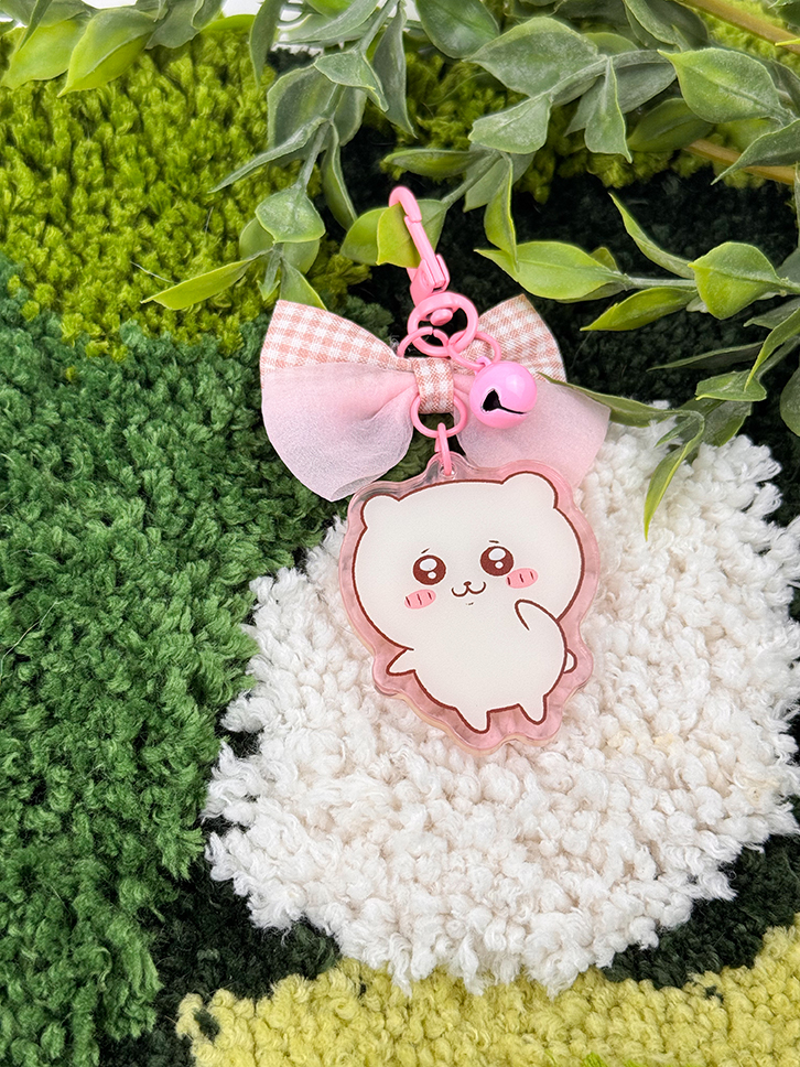 A Pink Hamster ~ Acrylic Keychain with a cartoon charm, pink bow, and pink bell rests on a textured green and white rug decorated with leafy plant accents.
