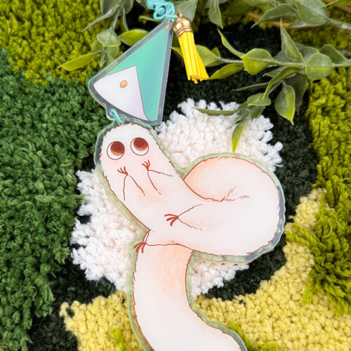 The Puffpuff ~ Acrylic Keychain features a cute, cartoonish white worm with big eyes, attached to a pastel geometric charm and yellow tassel, displayed on a green and yellow rug with artificial greenery.